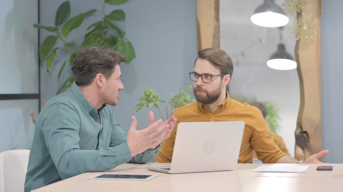 Two men sitting at a desk discussing work with a laptop in front.