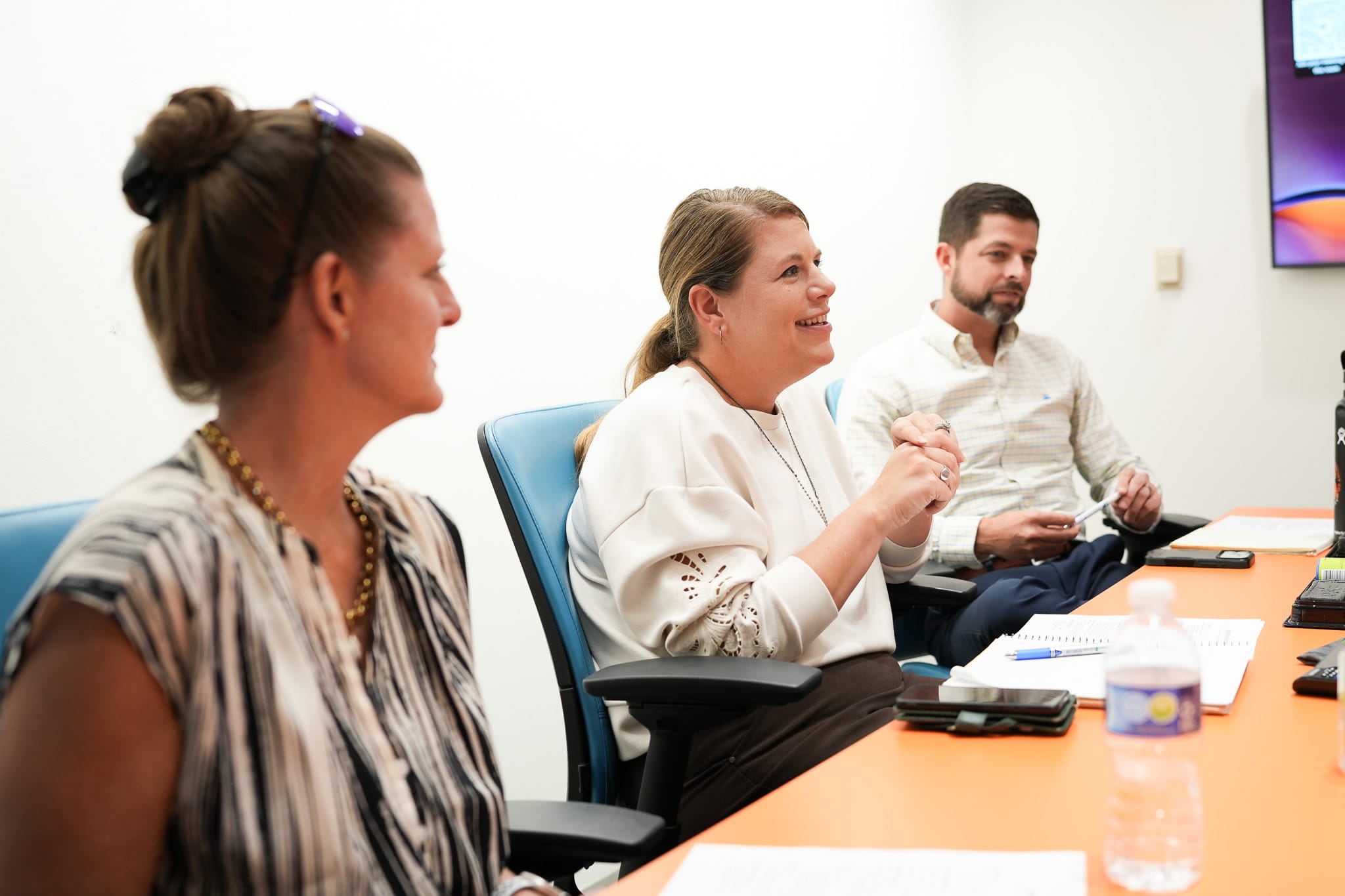 Three people sit around a table engaged in a meeting in a modern office setting.