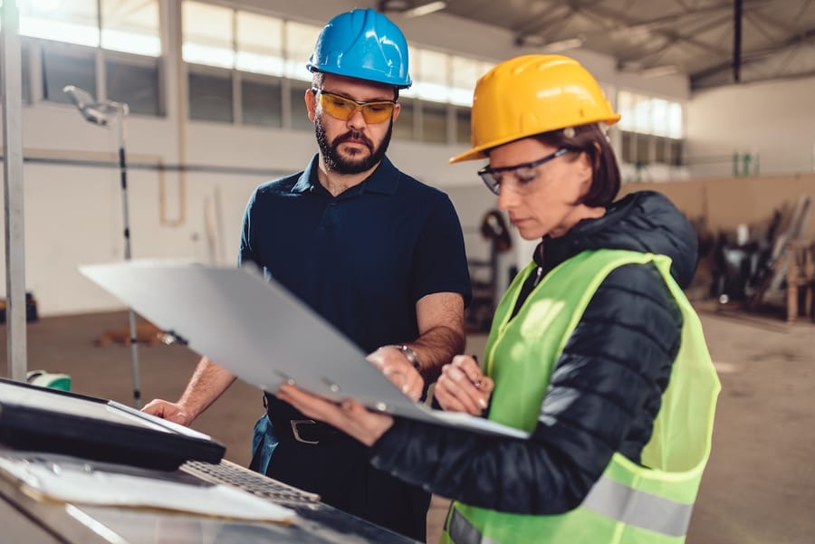 Two construction workers in hard hats reviewing plans on a clipboard in a warehouse