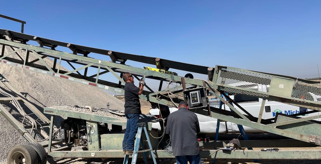 Two workers repairing an industrial conveyor system at an outdoor quarry or manufacturing facility