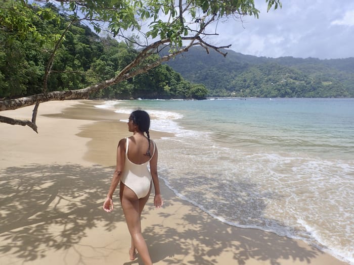 Woman in a white swimsuit walking along a deserted beach with trees and mountains.