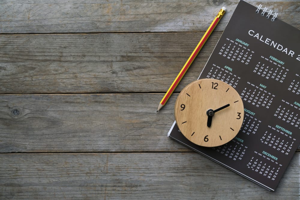 A pencil, wooden clock, and calendar lay on a wooden surface.